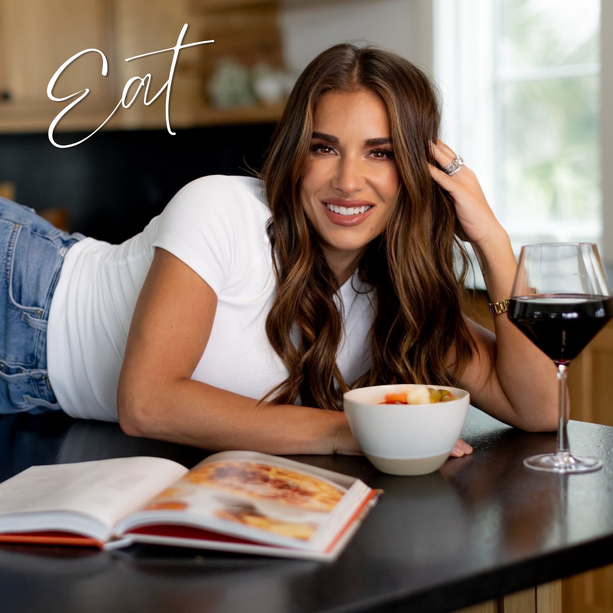 Jessie sitting at a table with a bowl of food, a glass of red wine, and an open book with words "Eat" on display