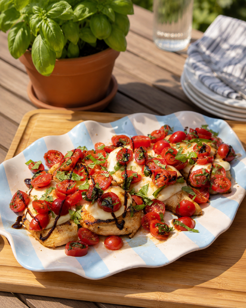 A scalloped ceramic platter holding grilled chicken topped with halved cherry tomatoes, melted cheese, fresh basil, and a drizzle of balsamic, set on a wooden board outdoors with a potted basil plant and stacked plates in the background.